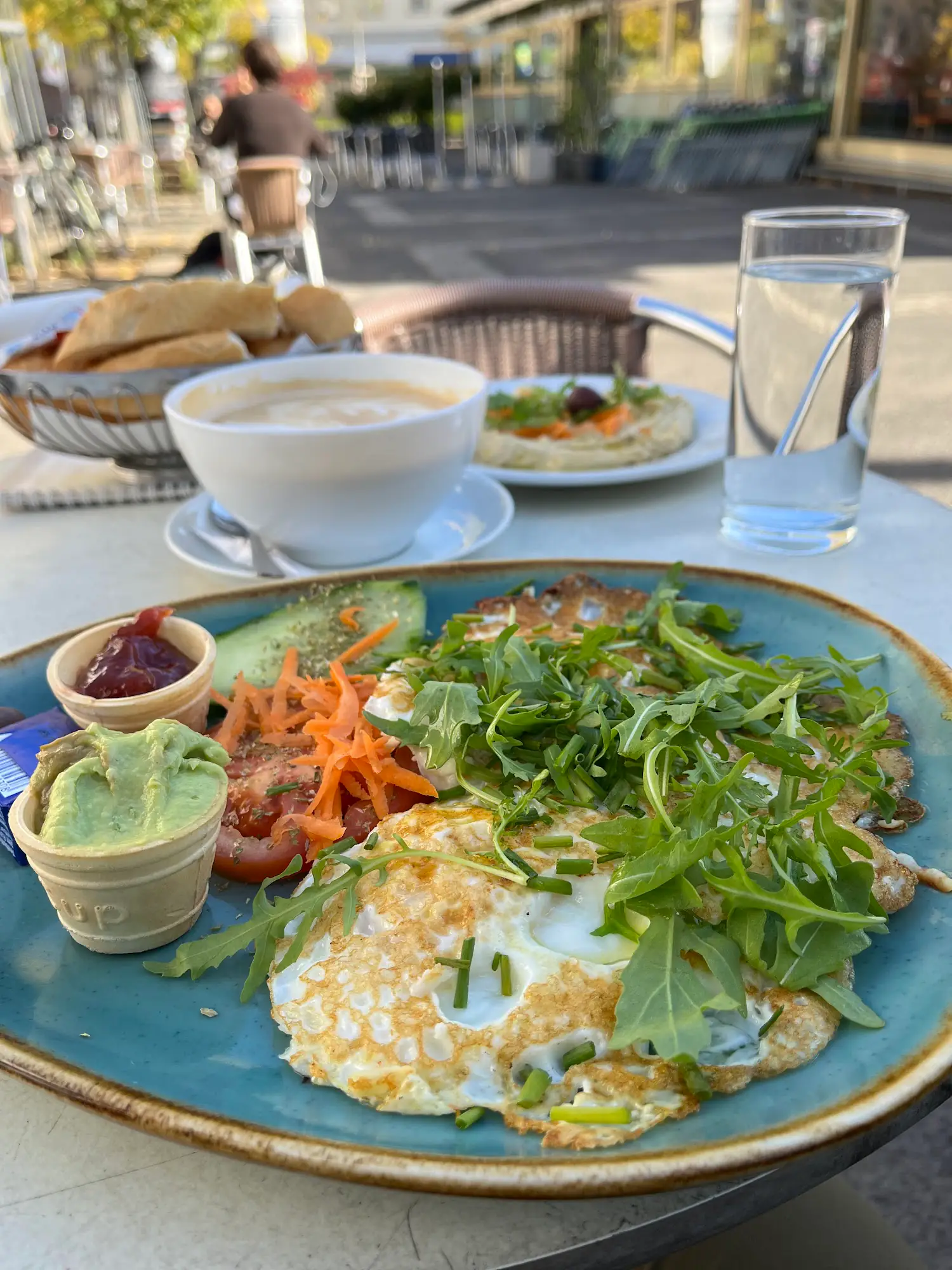 Frühstücksteller mit Spiegeleiern, Rucola, Dips, Brot und Kaffee auf einem Terrassentisch